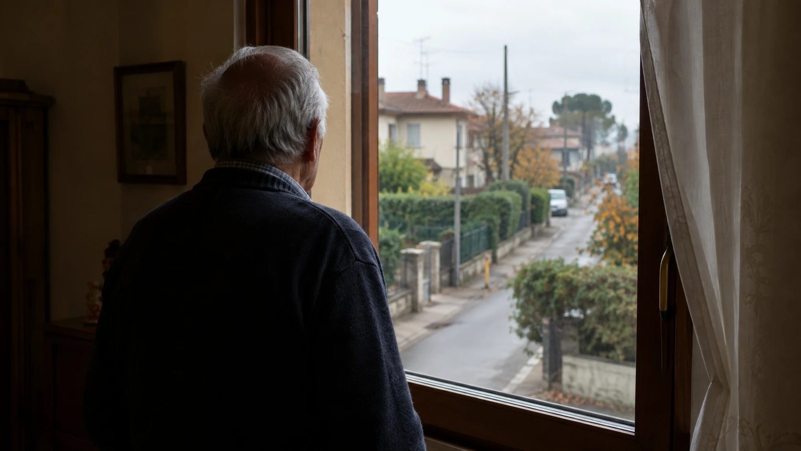 Un uomo anziano di spalle guarda fuori dalla finestra di casa, su una strada residenziale deserta